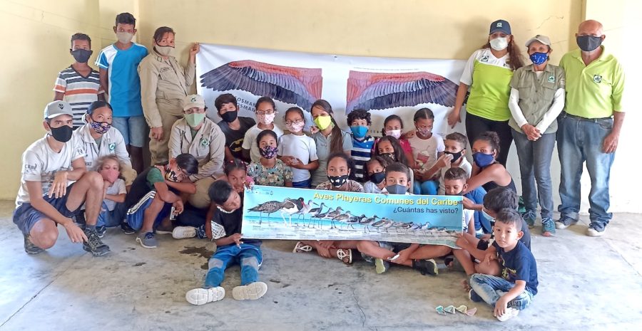 A group photo of young children sitting on the floor, while holding a shorebirds of the Caribbean poster. In the background two adults hold a banner with a pair of life-sized flamingo wings printed on it.