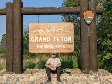 Chris is sitting in front of the Grand Teton National Park sign.