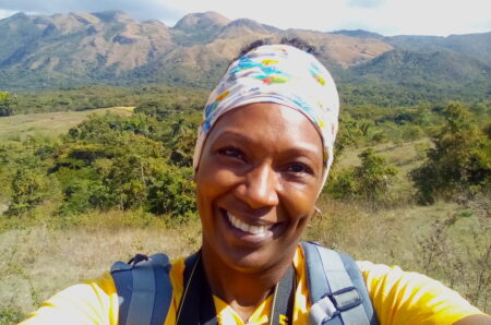 Selfie of a woman smiling at the camera, bag and binocular straps are partially visible. In the background are bare mountain ridges