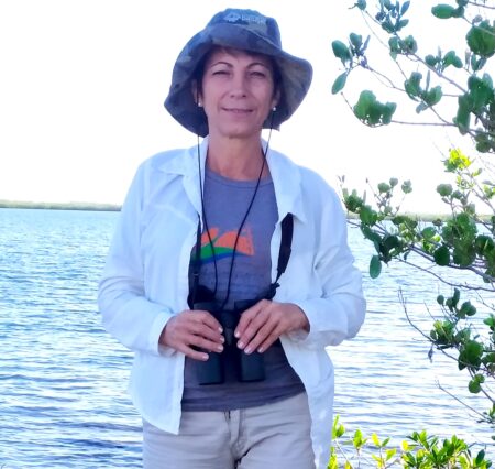 Photo of a woman holding binoculars. To the right there is a plant and in the background, a body of water. 