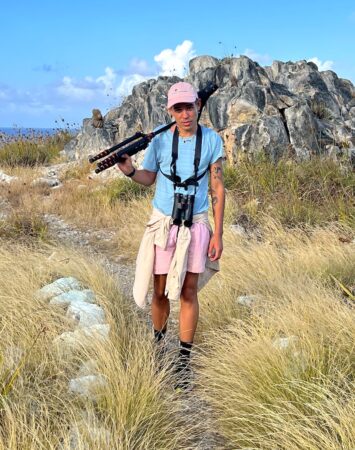 Photo of a man standing standing in front of a rocky outcrop with spotting scope over his right shoulder and binoculars around his neck. 