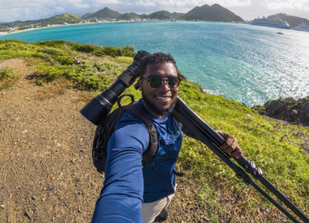 Selfie of a man wearing a blue long sleeve t-shirt. He is holding a camera with long lens and is attached to a tripod over his left shoulder. In the background, turquoise water and mountain ridges. 