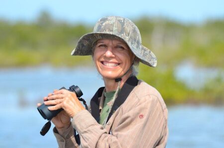 Photo of woman holding binoculars and smiling at camera. Body is turned away from camera but head is facing forward. 