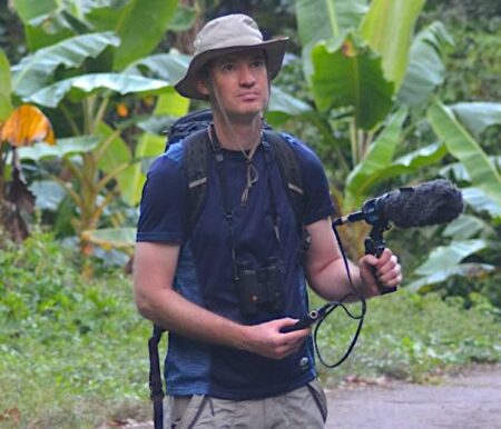 Photo of man holding a microphone in his left hand which is attached to a recorder in his right hand. He is wearing a hat and in the background there are plants. 