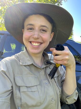 Selfie of woman holding a Puerto Rican Oriole in her left hand. Bird's head and tail can be clearly seen. 