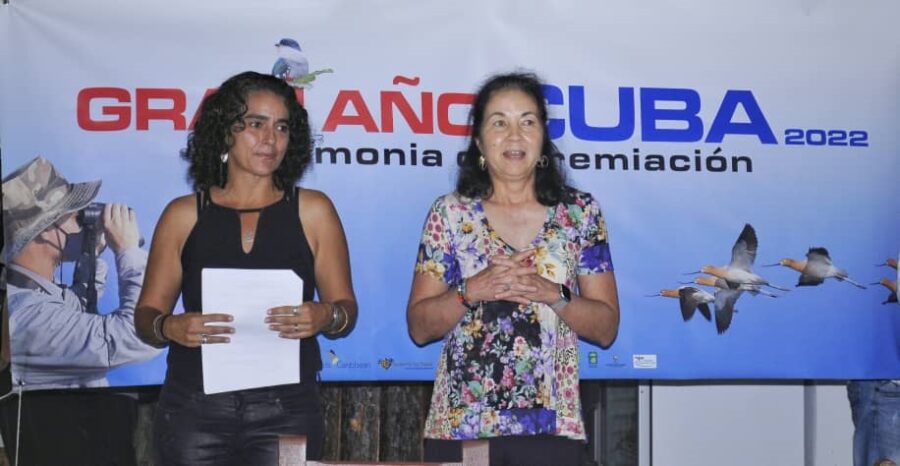 Photo of two women on stage facing forward and standing side by side. One woman is holding papers while the other makes a speech. The backdrop reads "Gran Ano Cuba" with a photo of a birder watching birds with binoculars to the left and birds flying on the right. 