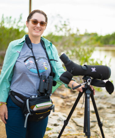 A woman standing next to a spotting scope. She is wearing a jacket, a pair of binoculars hangs from her neck, and a birding waist bag with just the top of a field guide visible. In the background there is a pond.