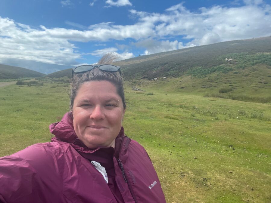 Selfie of a woman in a winter jacket outside. In the background there are green gently sloping hills. 