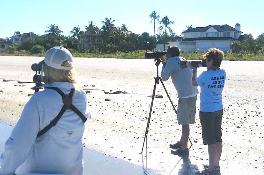 Photo of three people, one of which is looking through a spotting scope and the other two are looking through binoculars, on a beach in Florida. 