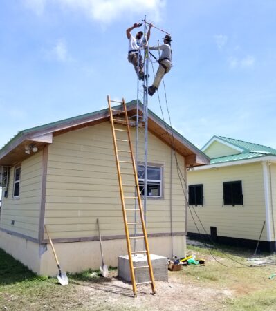 Photo of two men at the top of a Motus tower attaching an antenna. The tower is located behind a one storey building. To the left of the tower there is a lean to ladder.