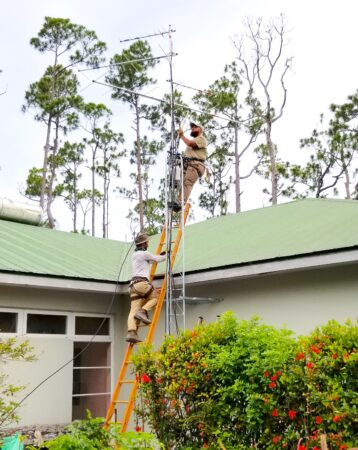 Photo of two men attaching an antenna to the Motus tower. One is standing on the tower while the other is climbing up the ladder.