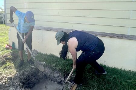 Photo of two women digging a hole in the ground behind a building.