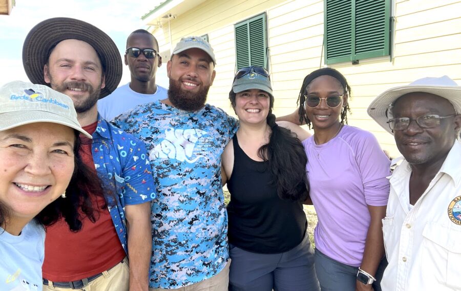 Photo (selfie) of group of 7 people standing in a line and smiling. In the background there is a one storey building.