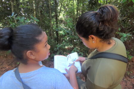 Two girls standing in a forested area. One is holding a book open and the other is pointing at a bird in the book.