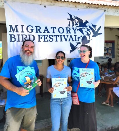 Group of three people- two women and one man, stand in front of a 'Migratory Bird Festival' sign while each holding a book.