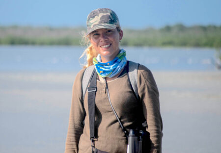 Photo of a woman wearing a hat and buff around her neck facing the camera and smiling. 