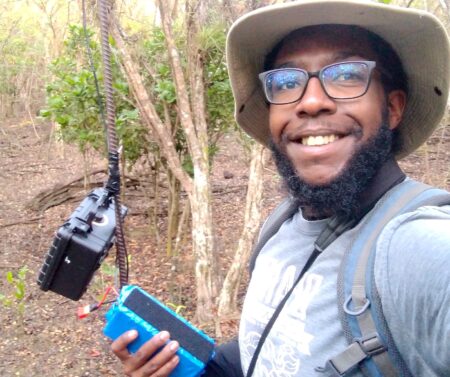 Selfie of a man in the forest holding a battery and next to him is a camera trap.