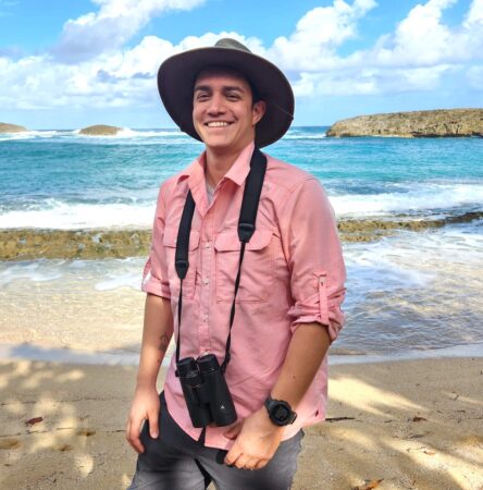 Photo of a man on a beach. He is facing forward with a binocular hanging from his neck, in the background is the ocean. 