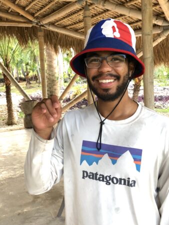 Photo of a man under a carrot shed facing the camera and smiling. He is holding a lizard in his hand.