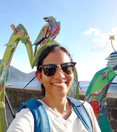 selfie of a woman wearing sunglasses and a bakcpack. In the background is a cut-out sign that spells Dominica.