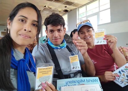 Selfie of three persons seated in the crowd- two female and one male- holding up their name tags and bird ID sheets to the camera.