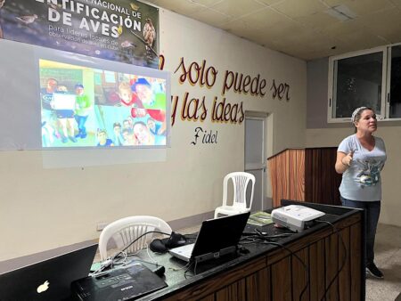 Photo of woman presenting, left of her is a desk with the projector and computer, and behind is a wall which the presentation is projected upon.