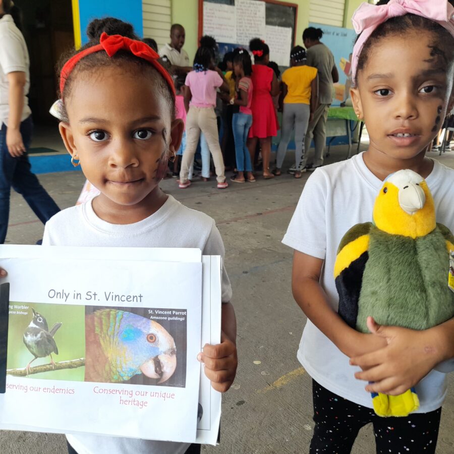 Two young girls, one holding a St Vincent Parrot plushie and the other holding printed photos of Whistling Warbler and St Vincent Parrot.