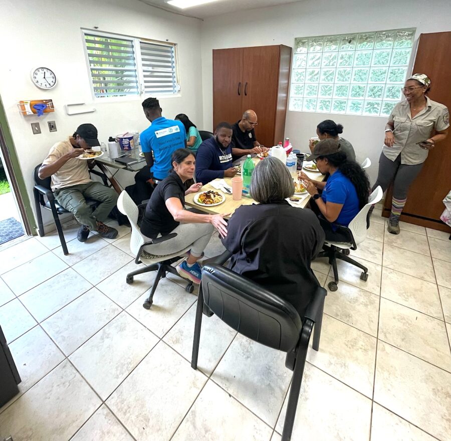 Photo of group of people sitting around two tables and having lunch together. 