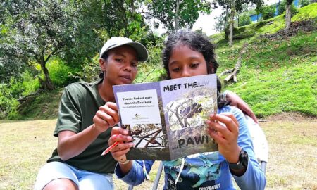 An adult woman and young make child both reading from a book titled "Meet the Pawi." They are sitting next to each other in a savanna. 