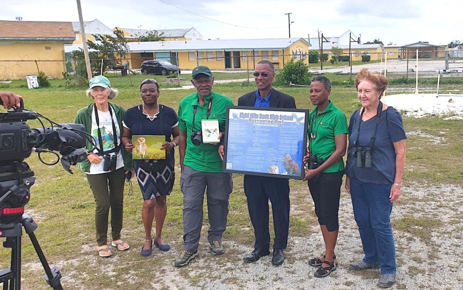 A group of seven people, likely members and staff of a Grand Bahama organization, stand outdoors being interviewed by a local TV station. From left to right, a person in a white t-shirt and green hat holds binoculars and a camera. Next to her, a woman in a dark dress holds a book or framed picture featuring an owl. The next two men wear green shirts, with one holding a camera and a book, and the other, in a suit jacket, holding a large framed poster titled "High Rock High School Burrowing Owl." To their right, a woman in a green shirt holds binoculars, and finally, another woman in a dark blue shirt also holds binoculars. They are all standing on a grassy area with some buildings visible in the background under a cloudy sky.