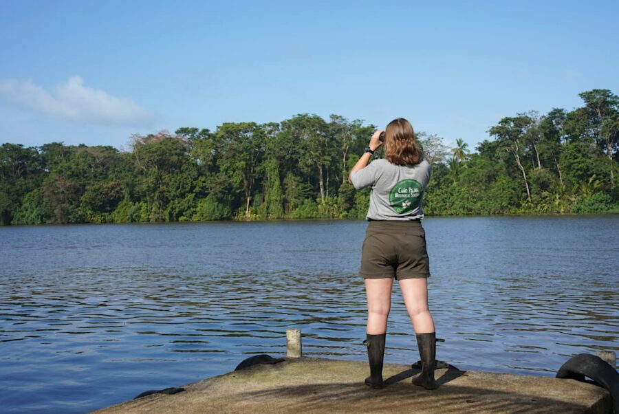 Photo of a woman standing on the bank of a river and is looking through binoculars to the other side of the river. 