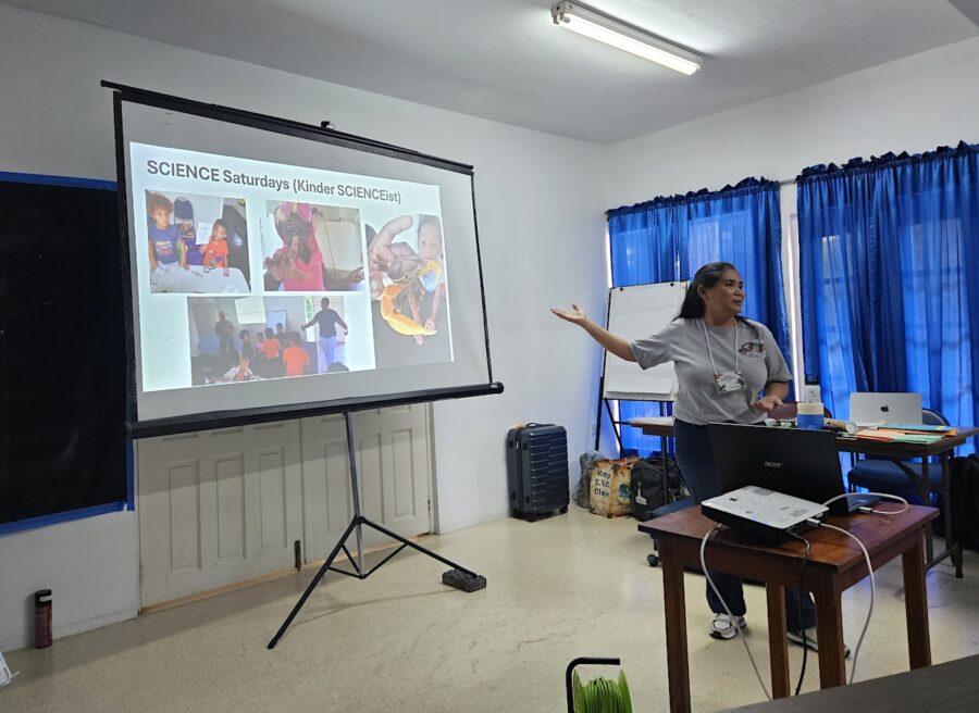 Photo of woman giving a presentation. She is standing in front of a projector screen and pointing to the screen which has the text SCIENCE Saturdays and photos of kids doing various science experiments. 