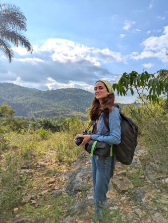 Photo of young woman standing in a clearing holding binoculars, in the background are mountain ridges. 