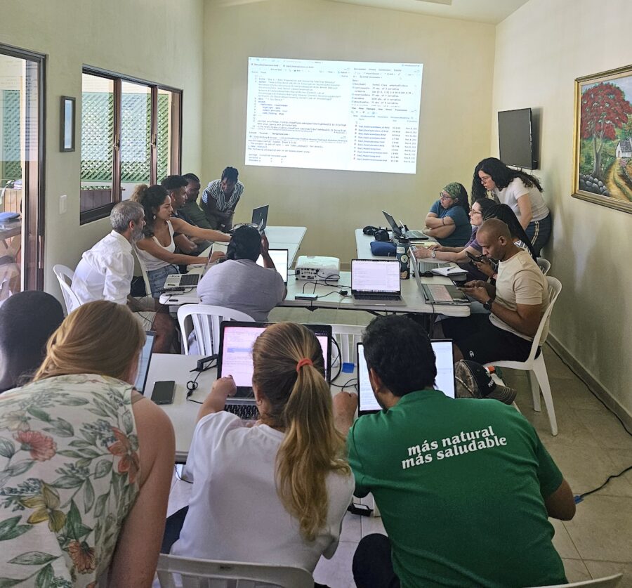 Photo of group of people in the room working on their laptops. Seating is U-shaped with one row to the back. Codes are projected on the wall in front of them. 