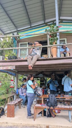 Photo of a young woman sitting on the balcony of the second story of a wooden house. The camera she is holding is resting on the railing on the balcony. 