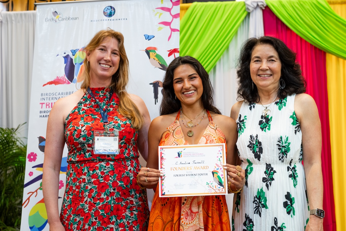 Photo of 3 women on a stage facing the camera. The woman in the middle is holding a certificate.