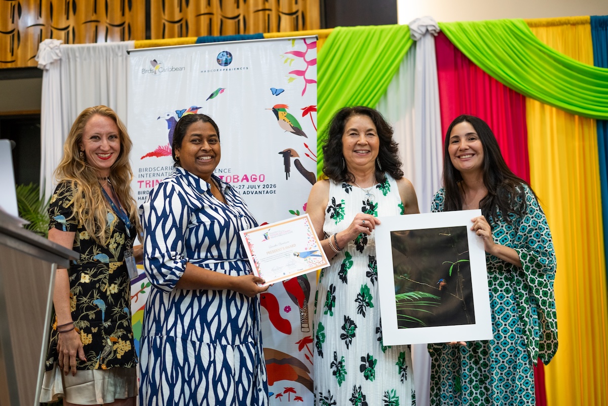Photo of four women on a stage facing the photographer. One woman is holding a certificate while another is holding a ready to frame photo of the Trinidad Motmot.