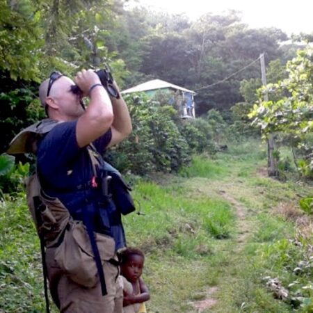 Photo of a man , taken from the side, he is looking up through binoculars with a small child standing next to him. The child is looking at the camera. They are standing in a bushy driveway with a house at the back. 
