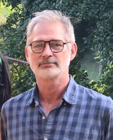 Portrait photo of a man wearing a blue plaid shirt and black glasses. 