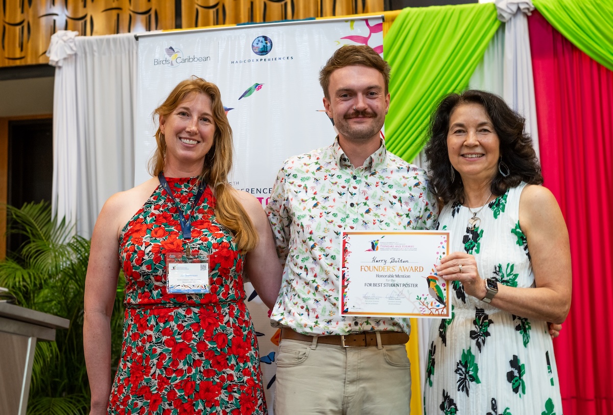 Photo of two women and one man standing on a stage facing the photographer. The man in the middle is holding a certificate.