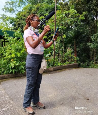 Photo of a woman holding a mic and phone to record bird sounds. In the background is a row of trees. 