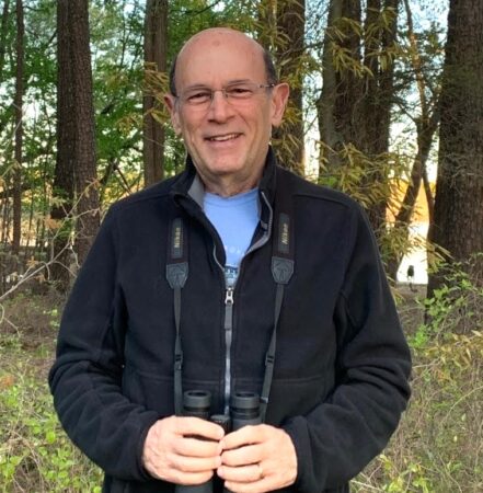 Photo of a man wearing a black jacket and blue t-shirt. A pair of binoculars is hanging from his neck which is he holding with both hands. He is looking directly at the camera. Behind him are a row of trees. 