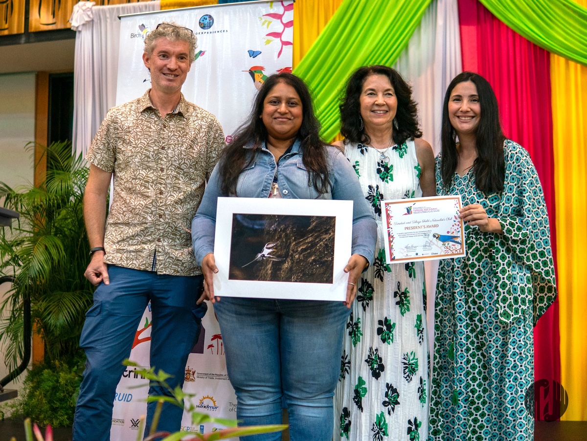 Photo of group of people- one man and three women-on a stage. One of the women is holding a ready to frame photo of a tropicbird while another woman is holding a certificate. They are all facing the photographer.