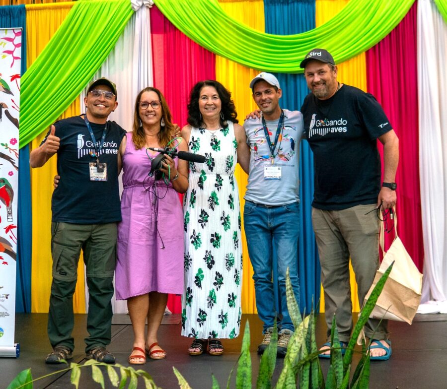 Photo of group of people standing on a stage. They are facing the photographer. From left to right: man (giving a thumbs up) next to him is a woman holding a microphone and headphones, next to them are a woman and two men. 