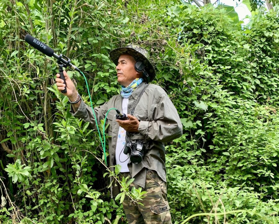 Photo of a man outdoors wearing headphones and holding up a microphone to record sounds. 